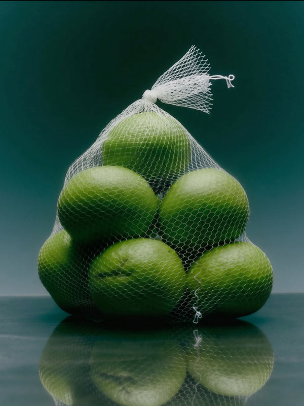 A bunch of green limes packed inside a white mesh bag, set against a dark teal background with a reflective surface below. Lime Salt